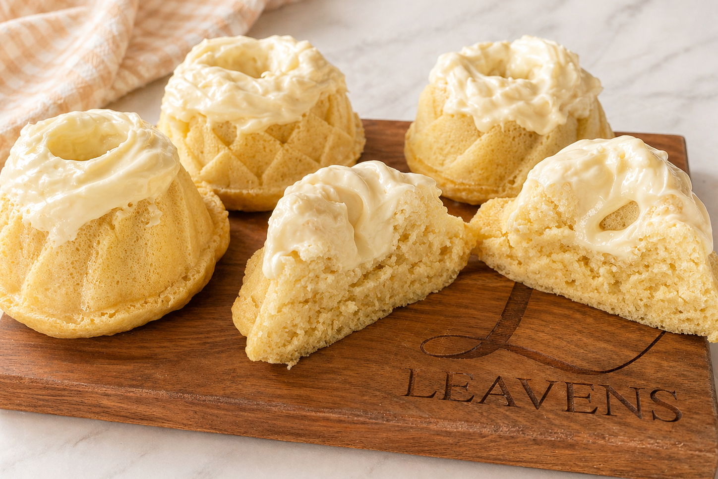 Four light yellow rolls made in different bundt-style pans. The rolls are topped with a white frosting and rest on a wooden cutting board. One roll is cut in half to show a fluffy, crumbling bread texture and the frosting pooling into the center hole.
