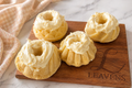 Four bundt shaped rolls topped with a whipped, white frosting. The rolls are placed on a wooden cutting board, which is resting on a marble counter with a kitchen cloth next to it.