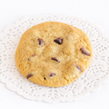 A large, round cookie with a soft, chewy texture, golden coloring, and several chocolate chips melted throughout it. The cookie is placed on a white doily.