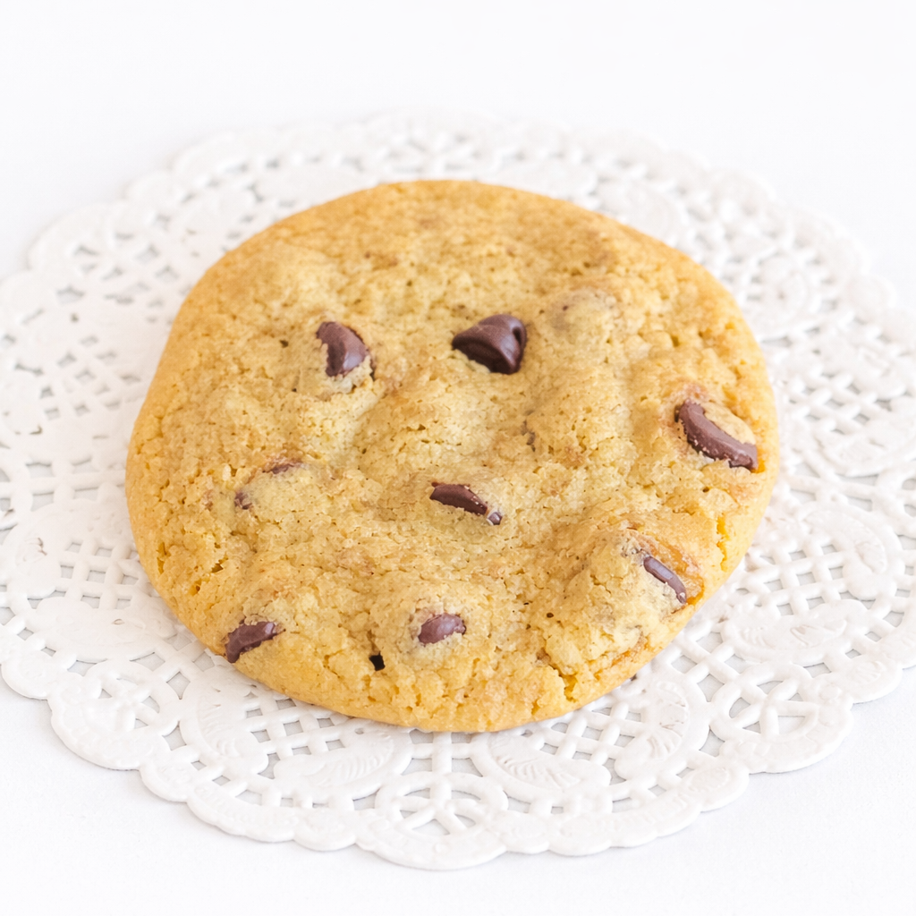 A large, round cookie with a soft, chewy texture, golden coloring, and several chocolate chips melted throughout it. The cookie is placed on a white doily.